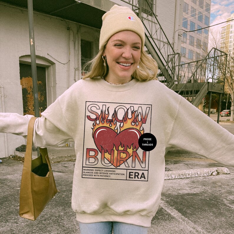 May include: A beige sweatshirt with a graphic print of a heart with flames and the text "SLOW BURN" in black. The text "PROSE + THREADS" is in a small black box in the upper right corner of the graphic. The text "ERA" is in the lower right corner of the graphic. The text "WARNING: EXPECT LINGERING GLANCES AND INTENSE ANTICIPATION PROCEED WITH PATIENCE" is in a small black box below the graphic.