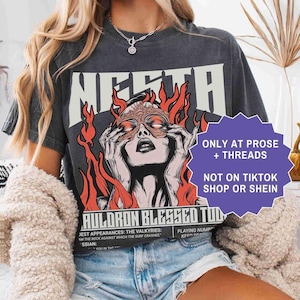May include: Dark gray t-shirt featuring a graphic of a woman with flames and the text "MERTA". The shirt also has the text "AULORON BLESSED TOD". The shirt is from Prose + Threads and not on TikTok Shop or Shein.