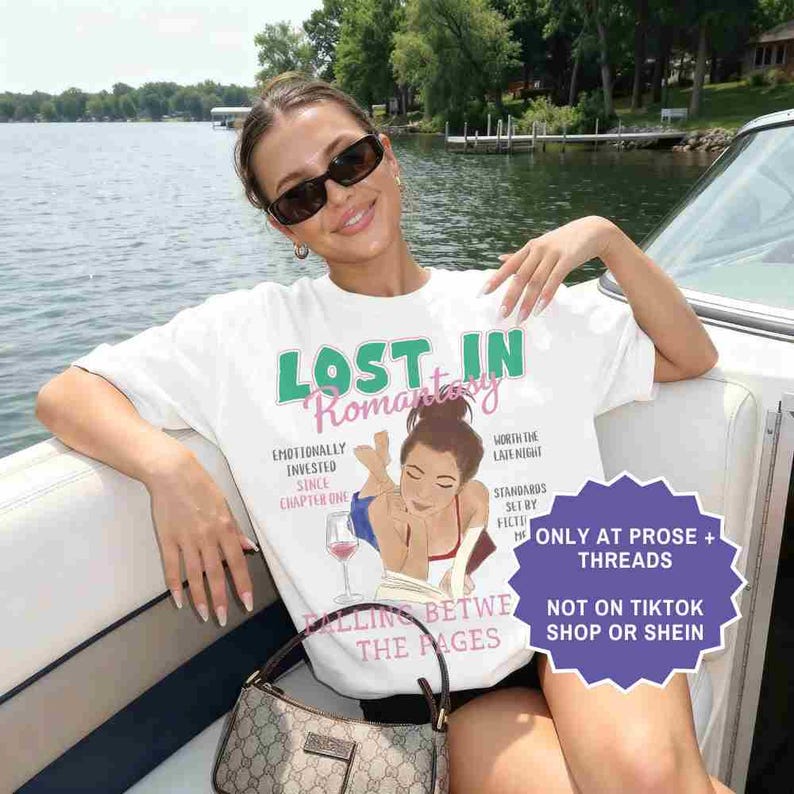 May include: White t-shirt with the text "LOST IN Romantasy" in green and pink. The shirt features an illustration of a woman reading, a wine glass, and additional text. A small purse is visible.