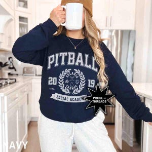 May include: Navy blue long-sleeved sweatshirt with a white graphic print of a wreath with the text "Solarbia Champs 2019" and "Zodiac Academy" below. The text "Pitball" is printed above the wreath.