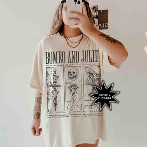 May include: Cream-colored t-shirt featuring the text "ROMEO AND JULIE" with illustrations of a sword, skull, rose, and wings. The shirt also includes the text "Verona" and a quote from William Shakespeare.