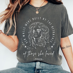 May include: Dark gray t-shirt featuring a white line drawing of Medusa with text that reads "HER WALLS WERE BUILT BY THE BETRAYALS of those she loved - MEDUSA-". The shirt is a crew neck and has short sleeves.
