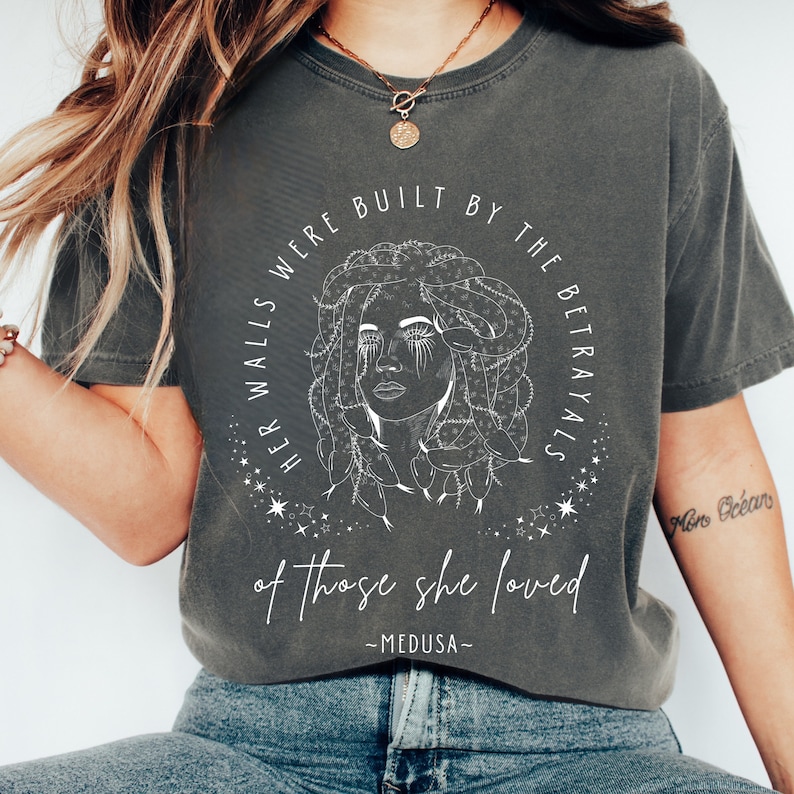 May include: Dark gray t-shirt featuring a white line drawing of Medusa with text that reads "HER WALLS WERE BUILT BY THE BETRAYALS of those she loved - MEDUSA-". The shirt is a crew neck and has short sleeves.