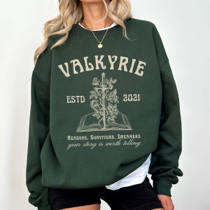 Może przedstawiać: Zielony sweter z nadrukiem "VALKYRIE ESTD 2021" i grafiką miecza w książce z r&oacute;żami. Tekst pod grafiką brzmi "Readers. Survivors. Dreamers your story is worth telling."