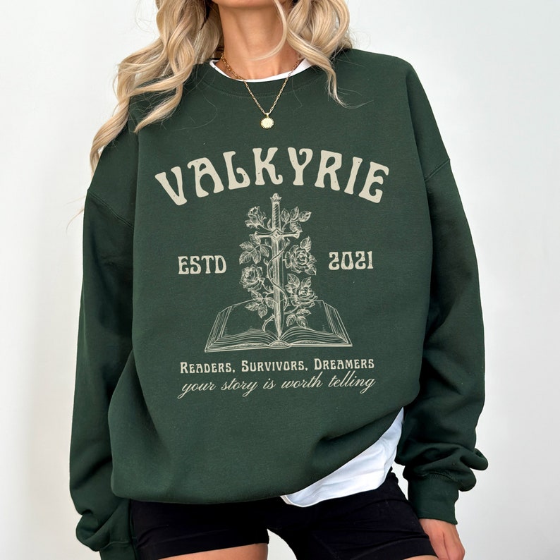 Może przedstawiać: Zielony sweter z nadrukiem "VALKYRIE ESTD 2021" i grafiką miecza w książce z r&oacute;żami. Tekst pod grafiką brzmi "Readers. Survivors. Dreamers your story is worth telling."
