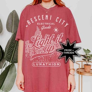 May include: A maroon t-shirt with a white graphic design that says "Crescent City Electrical Goods Light it Up Lunathion" with a star and a cityscape in the background. A black star with the text "Prose + Threads" is in the lower right corner.