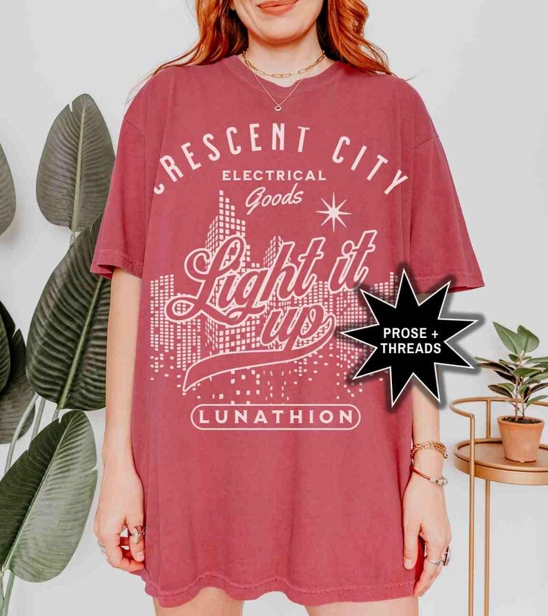 May include: A maroon t-shirt with a white graphic design that says "Crescent City Electrical Goods Light it Up Lunathion" with a star and a cityscape in the background. A black star with the text "Prose + Threads" is in the lower right corner.