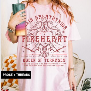 Pode incluir: Camiseta rosa claro com um gr&aacute;fico bord&ocirc;. O design inclui o texto "FIREHEART" e "QUEEN OF TERRASEN", junto com um cora&ccedil;&atilde;o, espadas e as palavras "RATTLE THE STARS". A marca "PROSE + THREADS" est&aacute; na parte inferior.