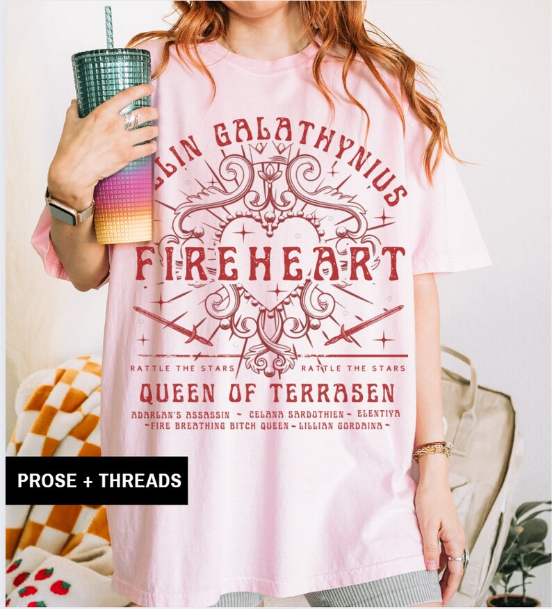 Pode incluir: Camiseta rosa claro com um gr&aacute;fico bord&ocirc;. O design inclui o texto "FIREHEART" e "QUEEN OF TERRASEN", junto com um cora&ccedil;&atilde;o, espadas e as palavras "RATTLE THE STARS". A marca "PROSE + THREADS" est&aacute; na parte inferior.