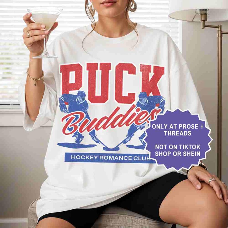 May include: White t-shirt with the words "PUCK Buddies" in red and blue. The shirt features two hockey players and the text "HOCKEY ROMANCE CLUB". A purple sticker says "ONLY AT PROSE + THREADS NOT ON TIKTOK SHOP OR SHEIN".