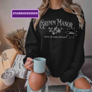 May include: Black crew neck sweatshirt with the text "Grimm Manor Home of Dark Romance" in white script. The sweatshirt also features a floral design and the words "Prose + Threads" in a starburst. The word "Embroidered" is in a purple box.