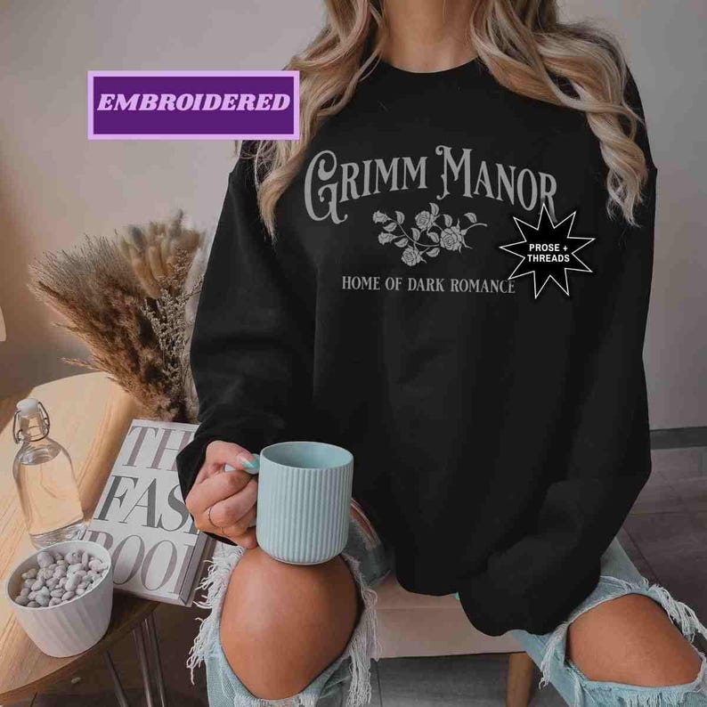 May include: Black crew neck sweatshirt with the text "Grimm Manor Home of Dark Romance" in white script. The sweatshirt also features a floral design and the words "Prose + Threads" in a starburst. The word "Embroidered" is in a purple box.