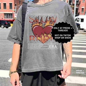 May include: Gray t-shirt with a graphic of a heart on fire, the words "SLOW BURN ERA" are printed above and below the heart. The shirt also has text that reads "WARNING: EXPECT LINGERING GLANCES AND INTENSE ANTICIPATION PROCEED WITH PATIENCE."