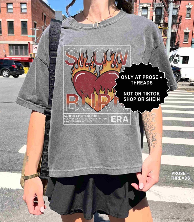 May include: Gray t-shirt with a graphic of a heart on fire, the words "SLOW BURN ERA" are printed above and below the heart. The shirt also has text that reads "WARNING: EXPECT LINGERING GLANCES AND INTENSE ANTICIPATION PROCEED WITH PATIENCE."