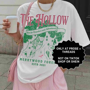 Pu&ograve; includere: T-shirt bianco con un design grafico con la scritta "THE HOLLOW" in rosa e verde, insieme a "Inn for Travelers and Adventurers" e "MERRYWOOD FORES ESTD 2021". La maglietta &egrave; abbinata a pantaloncini a righe rosa e bianche.
