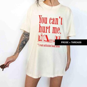 May include: A white t-shirt with the text "You can't hurt me." in red. Below the text is a graphic of a bookshelf with books. The text "*I read unfinished book series" is below the graphic.