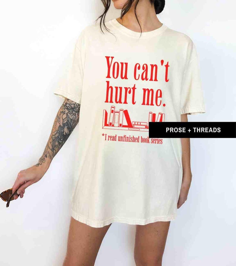 May include: A white t-shirt with the text "You can't hurt me." in red. Below the text is a graphic of a bookshelf with books. The text "*I read unfinished book series" is below the graphic.