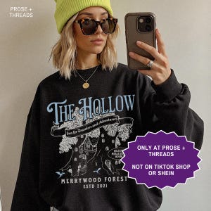 May include: Black sweatshirt with the text "THE HOLLOW" in light blue, featuring a treehouse graphic and the words "Inn for Travelers and Adventurers." The sweatshirt also has the text "MERRYWOOD FOREST ESTD 2021."