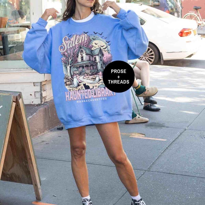 May include: A light blue sweatshirt with a graphic print of a house, trees, and ghosts. The text "Salem Haunted Library Massachusetts" is printed on the sweatshirt.