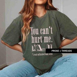 May include: A green t-shirt with the text "You can't hurt me." and a graphic of a bookshelf with books. The text below the graphic says "*I read unfinished book series". The text "PROSE + THREADS" is in the bottom right corner of the shirt.