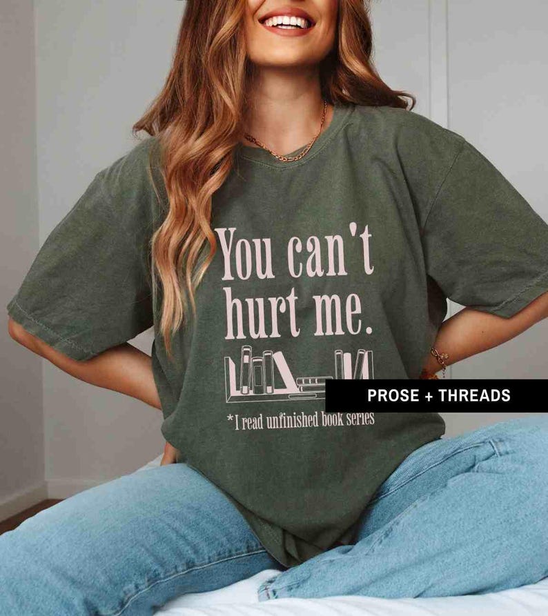 May include: A green t-shirt with the text "You can't hurt me." and a graphic of a bookshelf with books. The text below the graphic says "*I read unfinished book series". The text "PROSE + THREADS" is in the bottom right corner of the shirt.