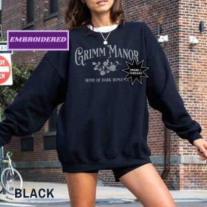 May include: A black sweatshirt featuring the text "Grimm Manor Home of Dark Romance" in silver. The sweatshirt has a crew neck and long sleeves. The word "Embroidered" is in a purple box. The word "Black" is in a black box in the lower left corner.