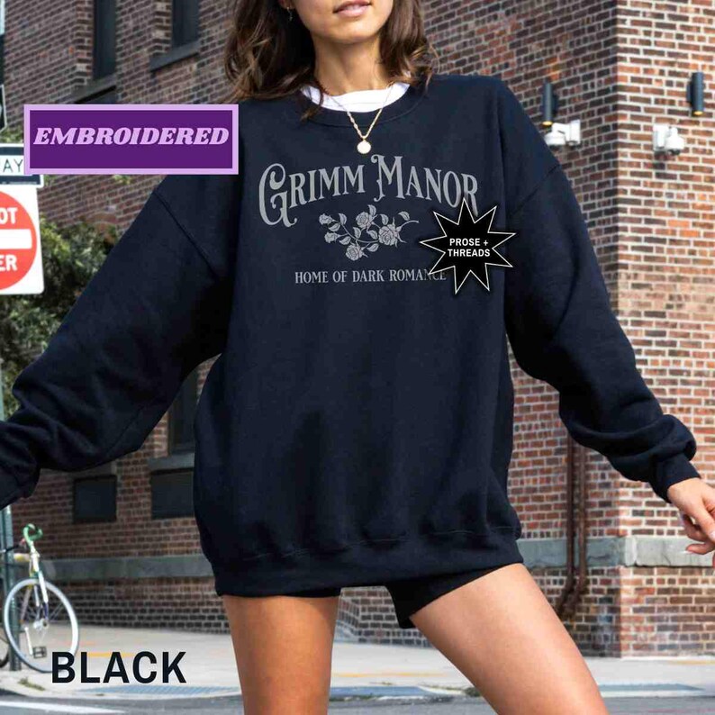 May include: A black sweatshirt featuring the text "Grimm Manor Home of Dark Romance" in silver. The sweatshirt has a crew neck and long sleeves. The word "Embroidered" is in a purple box. The word "Black" is in a black box in the lower left corner.