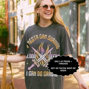May include: Dark gray t-shirt with the text "NESTA CAN SURVIVE" in a circular design, featuring crossed swords and flames. The shirt also includes the text "I CAN DO CARD". The shirt is worn by a person wearing sunglasses.