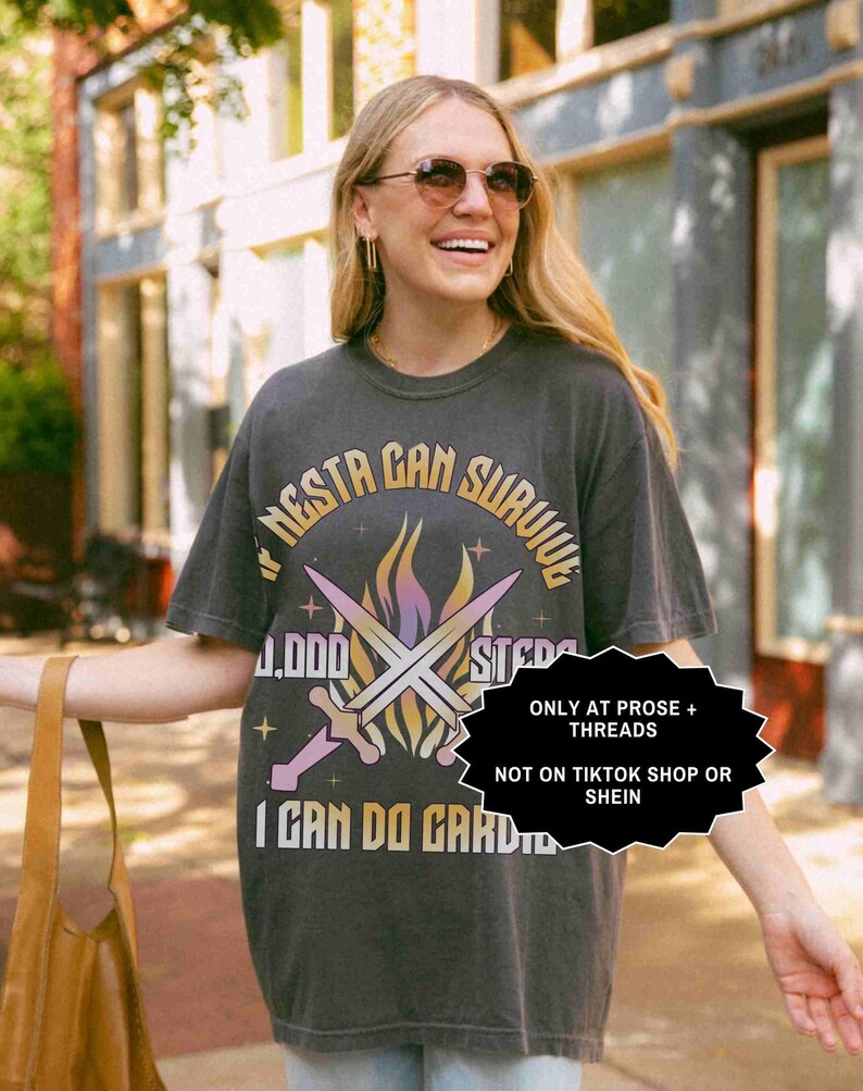 May include: Dark gray t-shirt with the text "NESTA CAN SURVIVE" in a circular design, featuring crossed swords and flames. The shirt also includes the text "I CAN DO CARD". The shirt is worn by a person wearing sunglasses.