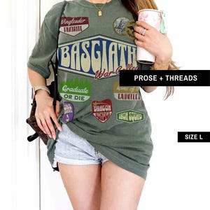 May include: Green oversized t-shirt with a vintage-style graphic print of "BASGIATH War College" and various patches, including "Vingleader Favorite", "Graduate or Die", "Dragon Bonder", "Iron Squad", and "Home of the Cauntile".