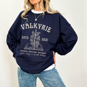 Może przedstawiać: Granatowa bluza z nadrukiem graficznym przedstawiającym książkę, miecz i r&oacute;że. Tekst "VALKYRIE ESTD 2021" jest nadrukowany nad grafiką. Tekst "READERS. SURVIVORS. DREAMERS your story is worth telling" jest nadrukowany pod grafiką.
