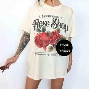 May include: A white oversized t-shirt with a vintage graphic design featuring a skull and crossbones, the text "Zade Meadows Rose Shop", two red roses, and the text "Open 24 Hours Discrete Delivery".