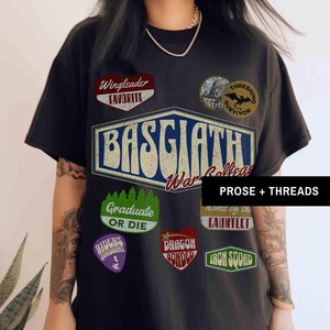 May include: Black t-shirt with a vintage-style graphic design featuring the text "Basgaathi War College" and various patches, including "Wingleader Favorite", "Thresing Survivor", "Graduate or Die", "Riders of the Radiant", "Dragon Bonded", "Home of the Gauntlet", and "Iron Squad".