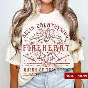 Pode incluir: Camiseta creme com um design gr&aacute;fico vermelho. O design apresenta as palavras "BELIN GALATHYNIUS FIREHEART" em uma fonte decorativa, juntamente com espadas e o texto "RATTLE THE STARS" e "QUEEN OF TER".