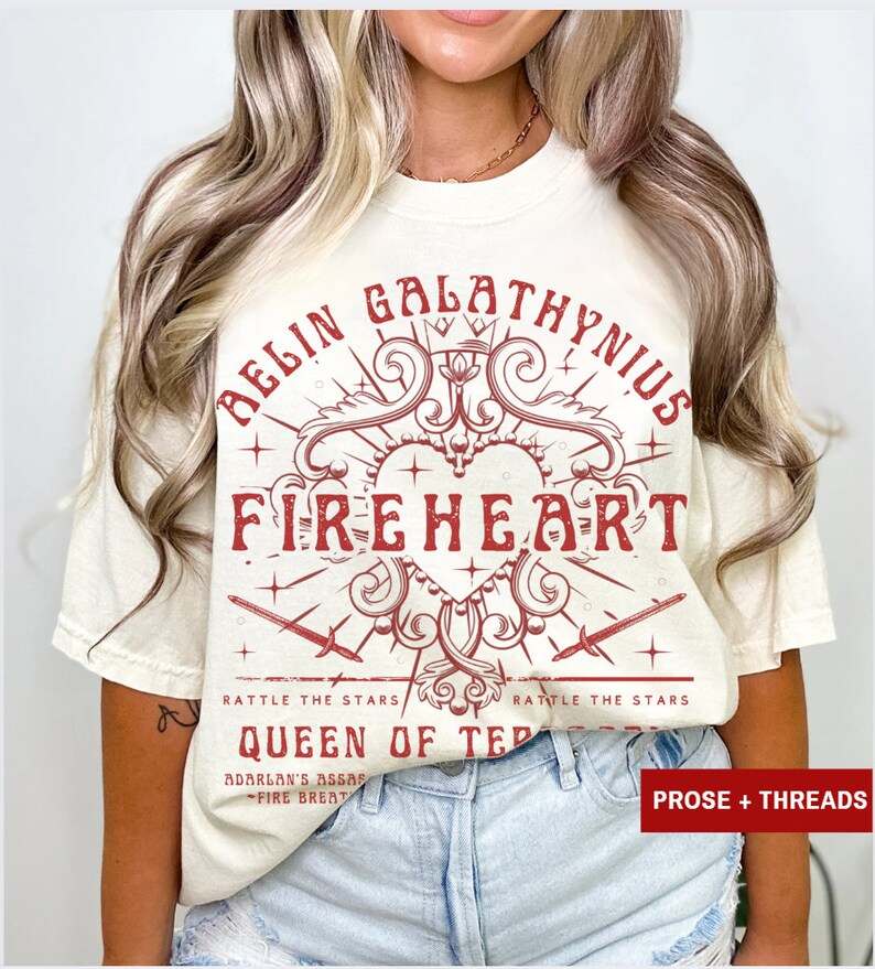 Pode incluir: Camiseta creme com um design gr&aacute;fico vermelho. O design apresenta as palavras "BELIN GALATHYNIUS FIREHEART" em uma fonte decorativa, juntamente com espadas e o texto "RATTLE THE STARS" e "QUEEN OF TER".