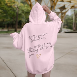 May include: A light pink hoodie with the text "To the person behind me. Life is tough my darling, but so are you" in black script, and a gold heart graphic. The hoodie has a hood and long sleeves.