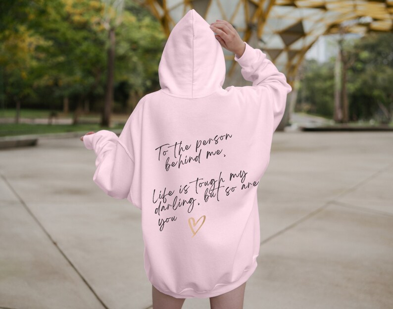 May include: A light pink hoodie with the text "To the person behind me. Life is tough my darling, but so are you" in black script, and a gold heart graphic. The hoodie has a hood and long sleeves.