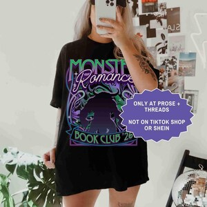Op de afbeelding: Zwart T-shirt met de tekst "Monster Romance Book Club '26" in een neon groen en paars ontwerp. Het shirt heeft ook een silhouet van een monster. Een zwarte baseball cap wordt gedragen.