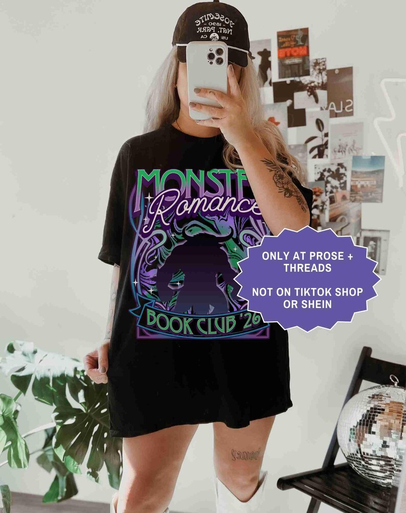 Op de afbeelding: Zwart T-shirt met de tekst "Monster Romance Book Club '26" in een neon groen en paars ontwerp. Het shirt heeft ook een silhouet van een monster. Een zwarte baseball cap wordt gedragen.