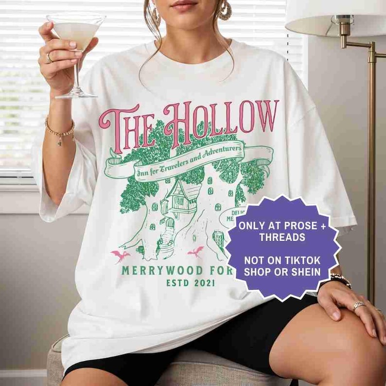 Pu&ograve; includere: T-shirt bianco con un design grafico con la scritta "THE HOLLOW" in rosa, con un'illustrazione di albero e casa verde. La maglietta include anche il testo "Inn for Travelers and Adventurers" e "MERRYWOOD FOR ESTD 2021".