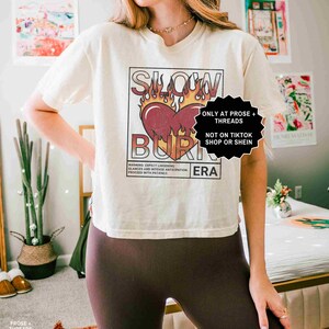 May include: Cream-colored t-shirt with a graphic design featuring a heart engulfed in flames, the words "SLOW BURN ERA" are printed above and below the heart. The shirt also has the text "WARNING: EXPECT LINGERING GLANCES AND INTENSE ANTICIPATION. PROCEED WITH PATIENCE."