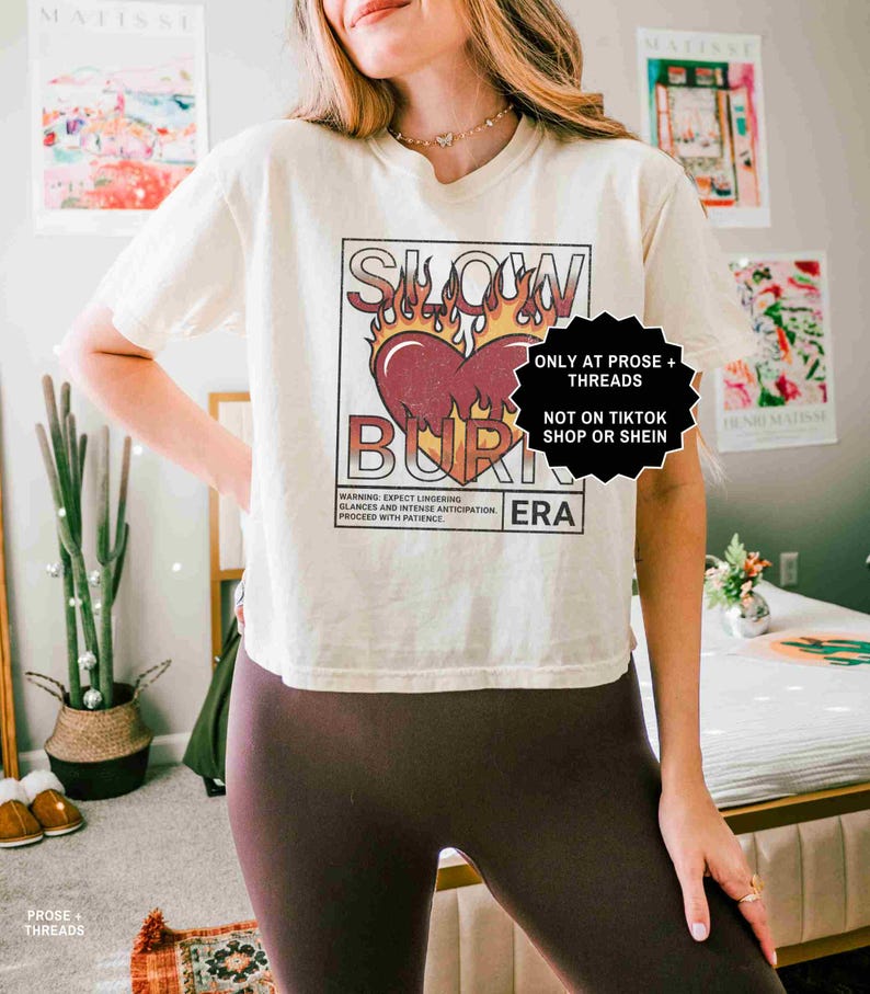 May include: Cream-colored t-shirt with a graphic design featuring a heart engulfed in flames, the words "SLOW BURN ERA" are printed above and below the heart. The shirt also has the text "WARNING: EXPECT LINGERING GLANCES AND INTENSE ANTICIPATION. PROCEED WITH PATIENCE."