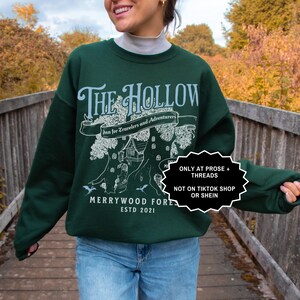 May include: A dark green sweatshirt featuring the words "The Hollow" in white, with a tree graphic and the text "Inn for Travelers and Adventurers". The sweatshirt also displays "Merrywood Forest ESTD 2021".