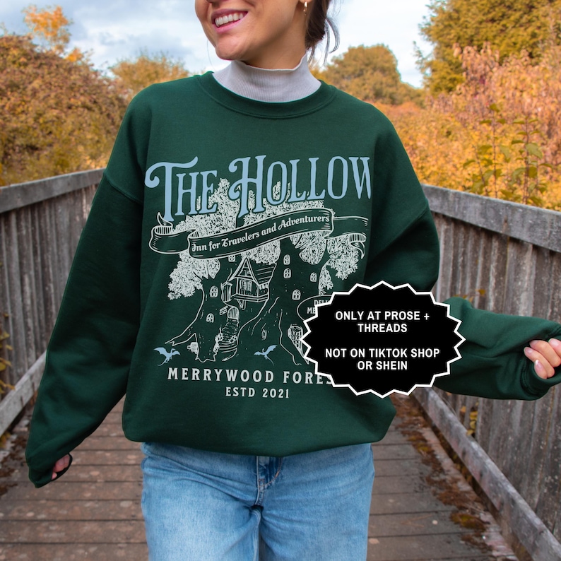 May include: A dark green sweatshirt featuring the words "The Hollow" in white, with a tree graphic and the text "Inn for Travelers and Adventurers". The sweatshirt also displays "Merrywood Forest ESTD 2021".