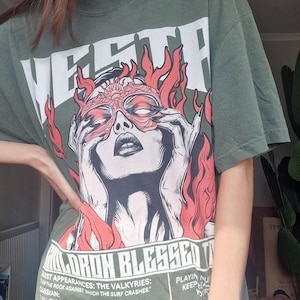 May include: Olive green graphic t-shirt featuring a stylized illustration of a woman with a mask and flames. The shirt has the word "VESTP" at the top and text at the bottom. A casual, oversized fit.