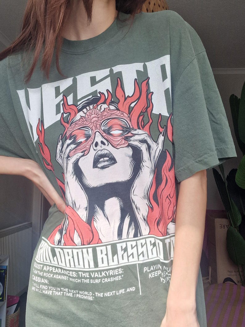 May include: Olive green graphic t-shirt featuring a stylized illustration of a woman with a mask and flames. The shirt has the word "VESTP" at the top and text at the bottom. A casual, oversized fit.