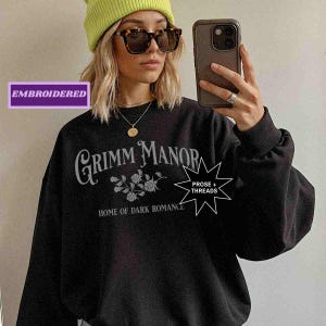 May include: Black sweatshirt featuring the text "Grimm Manor Home of Dark Romance" in a gothic font, with a floral design. The word "Embroidered" is in a purple rectangle. The sweatshirt is worn with a yellow beanie and sunglasses.