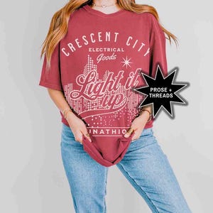 May include: A maroon oversized t-shirt with a white graphic design that says "Crescent City Electrical Goods Light it Up" with a cityscape graphic and a star. The graphic is in a retro style font.