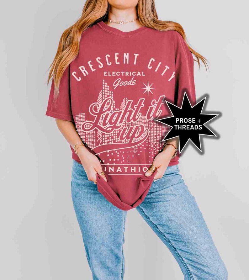 May include: A maroon oversized t-shirt with a white graphic design that says "Crescent City Electrical Goods Light it Up" with a cityscape graphic and a star. The graphic is in a retro style font.