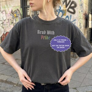Pu&ograve; includere: Una t-shirt a maniche corte grigio antracite con il testo Read With Pride in un carattere gotico, con la parola Pride visualizzata nei colori dell'arcobaleno. Un grafico viola a forma di stella include il testo ONLY AT PROSE + THREADS NOT ON TIKTOK SHOP OR SHEIN.